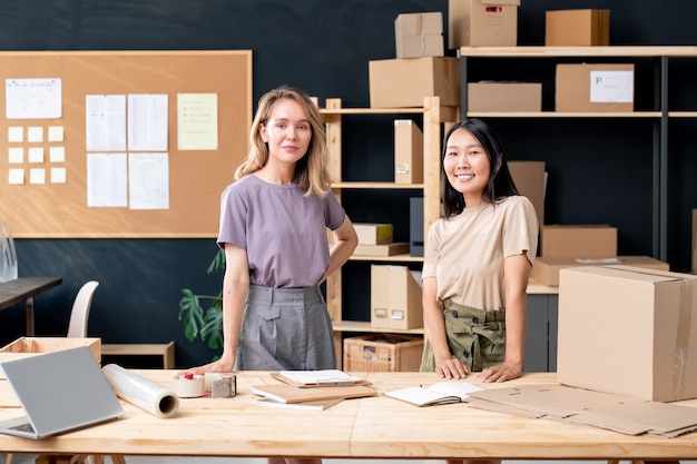 Jeunes jolies travailleuses interculturelles du bureau de la boutique en ligne debout près d'une table en bois tout en travaillant avec des commandes