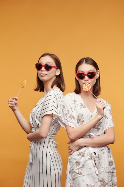 Jeunes jumeaux asiatiques magnifiques dans des robes élégantes et des lunettes de soleil élégantes ayant des sucettes tout en se tenant devant la caméra sur fond jaune