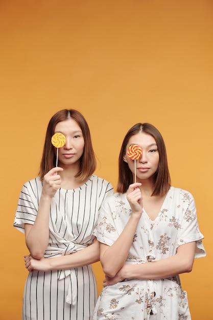 Jeunes jumeaux asiatiques magnifiques avec des sucettes par leurs yeux vous regardant tout en se tenant devant la caméra sur fond jaune