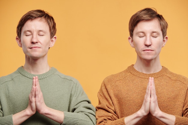 Jeunes jumeaux calmes et détendus en pulls se tenant la main dans des gestes de Namaste et méditant les yeux fermés sur fond orange