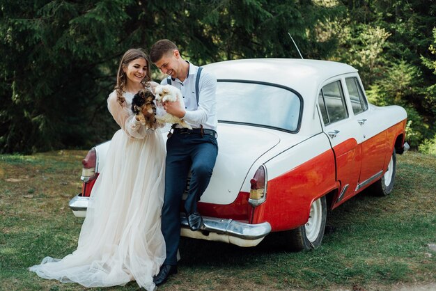 Les jeunes mariés aimants sont debout, se tenant la main, près d'une vieille voiture rétro blanche sur la nature en été. Portrait de mariage d'un marié élégant et souriant et d'une jolie mariée.