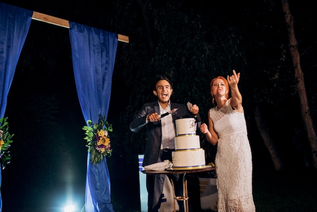 Les jeunes mariés coupent, rient et goûtent joyeusement le gâteau de mariage