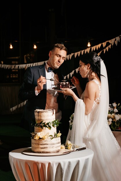 Les jeunes mariés coupent, rient et goûtent joyeusement le gâteau de mariage