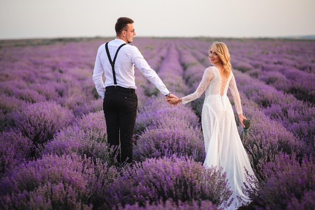 Les jeunes mariés habillés en style campagnard à pied se tenant la main le long d'un champ de lavande en fleurs