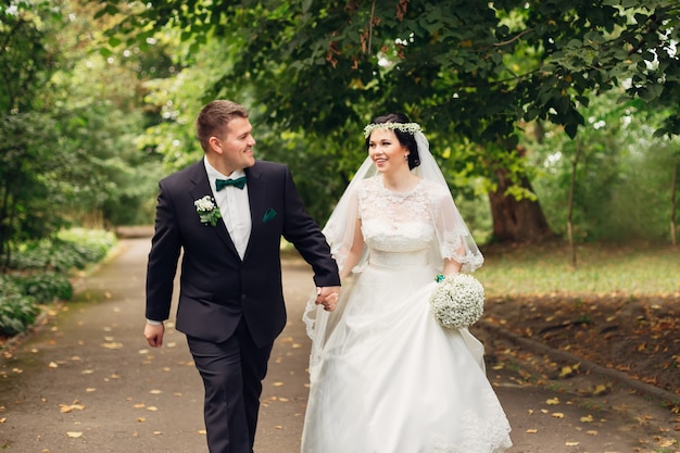 Les jeunes mariés marchent dans le parc et se regardent La mariée tient un bouquet de mariage