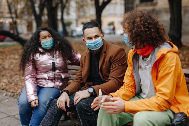 Jeunes multiethniques assis sur un banc en bois au parc de la ville et portant des masques médicaux pour se protéger du coronavirus. Concept de maladies infectieuses.