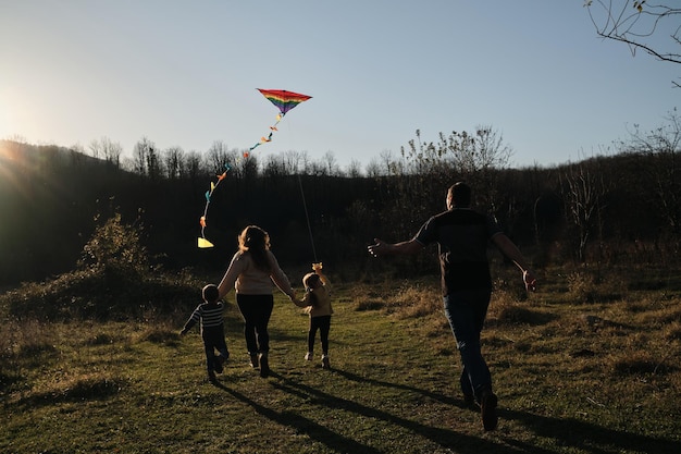 Les jeunes parents avec fils et fille traversent le champ au coucher du soleil et lancent un cerf-volant multicolore dans le ciel