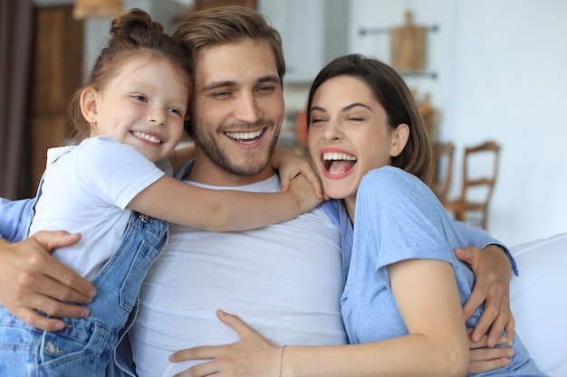 Jeunes parents positifs et sympathiques avec une petite fille souriante assise sur un canapé ensemble tout en se relaxant à la maison le week-end.