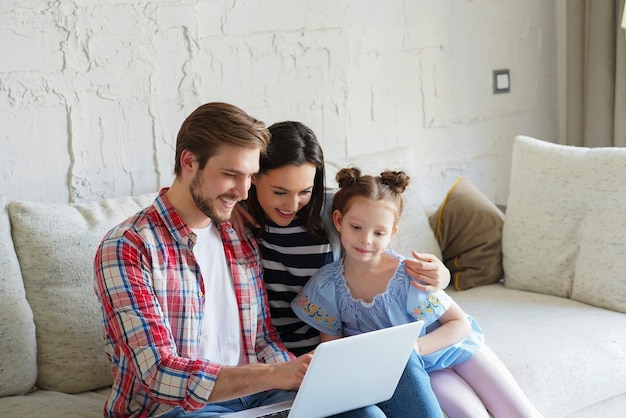 Jeunes parents positifs et sympathiques avec une petite fille souriante assise sur un canapé en train de regarder des photos sur un ordinateur portable, se relaxant à la maison le week-end.