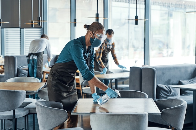 Jeunes Serveurs Masculins Occupés Dans Des Tables De Nettoyage De Vêtements De Travail Protecteurs Au Restaurant