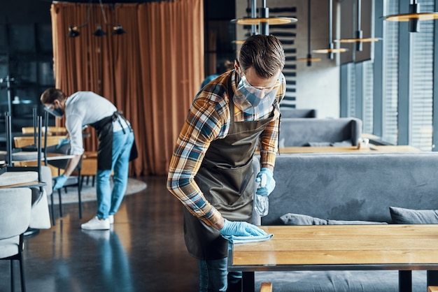 Jeunes serveurs masculins occupés dans des vêtements de travail protecteurs pulvérisant du désinfectant sur les tables du restaurant