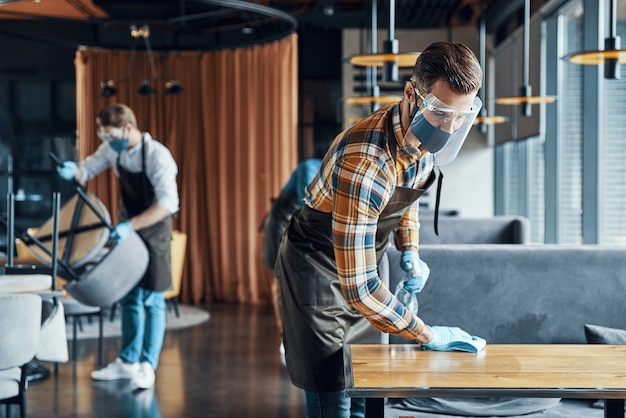 Photo jeunes serveurs masculins occupés dans des vêtements de travail protecteurs pulvérisant du désinfectant sur les tables du restaurant