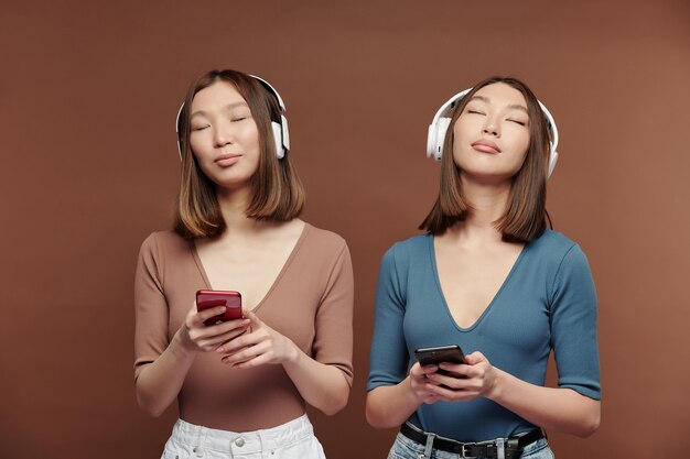 Jeunes sœurs jumelles asiatiques en tenue décontractée et écouteurs profitant d'une musique relaxante et utilisant des smartphones tout en se tenant devant la caméra