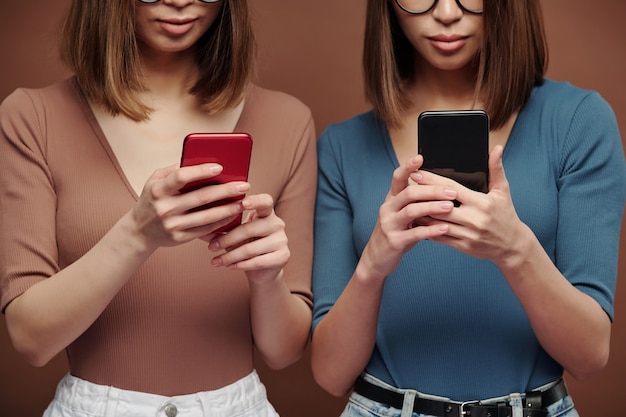 Jeunes soeurs jumelles brunes en tenue décontractée se tenant près les unes des autres et faisant défiler les smartphones devant la caméra sur fond marron