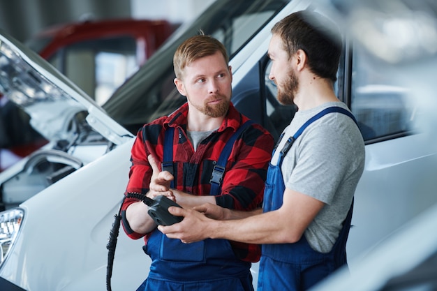 L'un des jeunes spécialistes de la réparation automobile pointant sur un appareil électronique entre les mains de son collègue pendant la conversation de travail