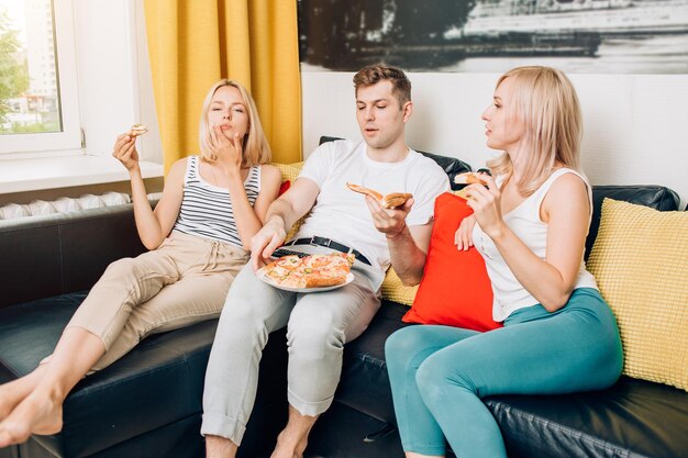 Des jeunes en tenue décontractée mangeant de la pizza, assis sur un canapé, parlant, regardant la télévision tout en se reposant à la maison. Week-end, loisirs, concept de nourriture malsaine.