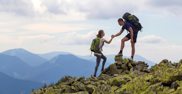 Les jeunes touristes avec des sacs à dos, un garçon athlétique aide une fille mince à affronter le sommet des montagnes rocheuses contre le ciel d'été et la scène de la chaîne de montagnes. Concept de tourisme, de voyage et de mode de vie sain.