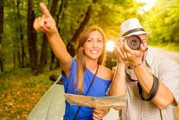 Jeunes voyageurs debout devant une voiture dans la forêt. Fille tenant la carte et pointant vers la destination. Guy prenant une photo avec un appareil photo numérique.