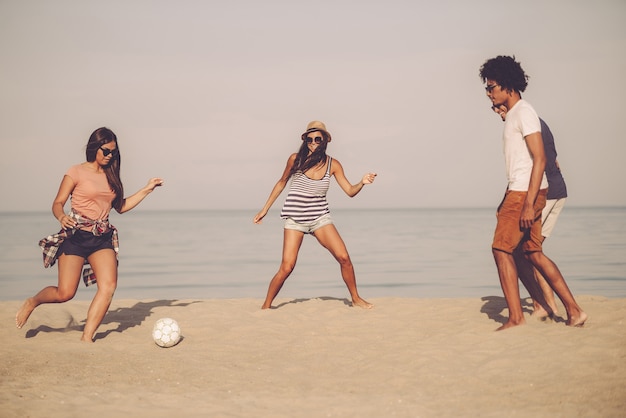 Jeux actifs avec des amis. Groupe de jeunes joyeux jouant avec un ballon de football sur la plage avec la mer en arrière-plan
