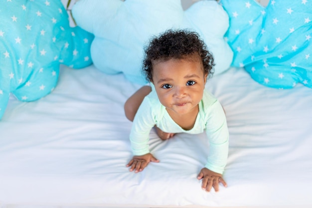 Joli bébé africain assis dans son lit à la maison avant d'aller se coucher sur un lit bleu et de rechercher le concept de sommeil sain et d'accessoires en coton pour la literie de bébé