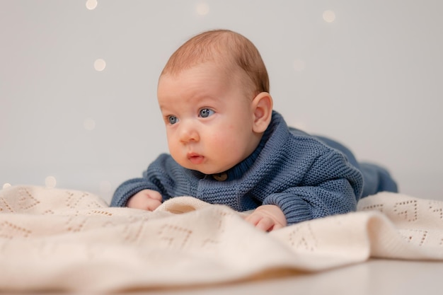 Joli bébé aux yeux bleus en combinaison tricotée bleue se trouve sur son ventre sur un enfant à carreaux gris apprend à