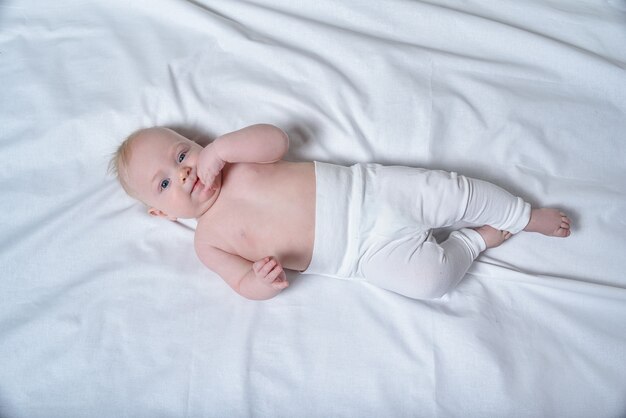 Joli bébé blond avec les doigts dans la bouche se trouve sur un drap blanc. Vue de dessus