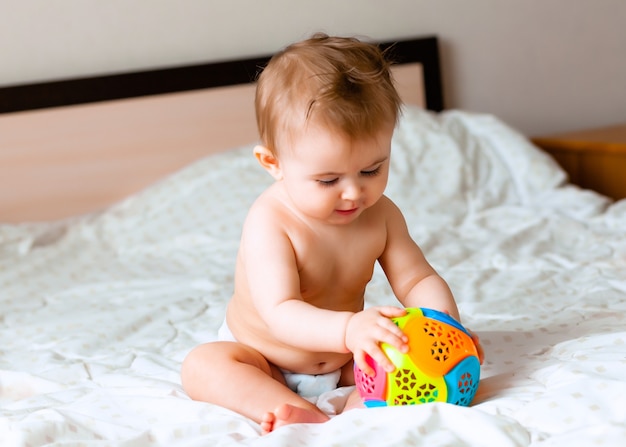 Joli bébé blond jouant avec un ballon assis sur le lit dans la chambre. heureux bébé de 6 mois jouant avec un ballon