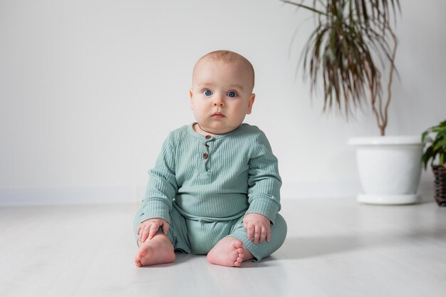 Joli bébé potelé dans une combinaison verte est assis sur le sol à côté d'une plante d'intérieur