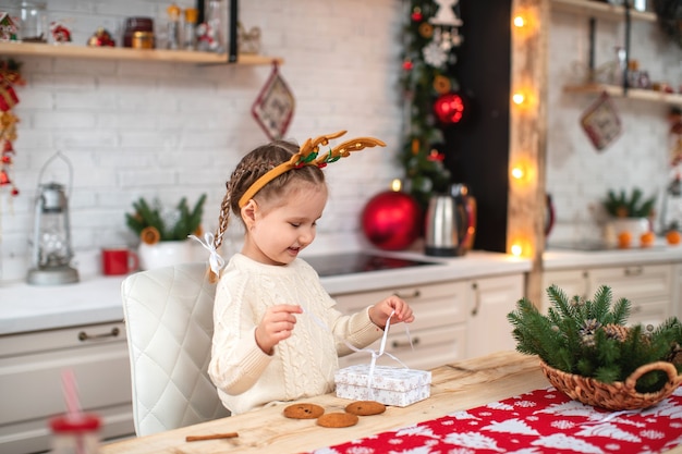 Joli bébé en pull léger et bandeau avec des cornes de renne assis à table de cuisine souriant ouvre la boîte-cadeau