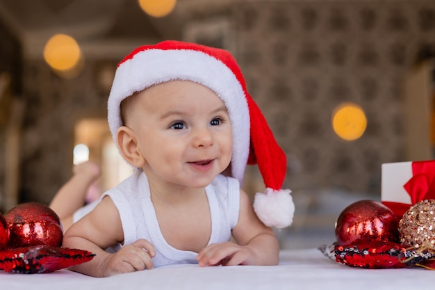 Joli bébé souriant dans un body blanc et un bonnet de Noel est allongé sur le ventre sur un drap blanc