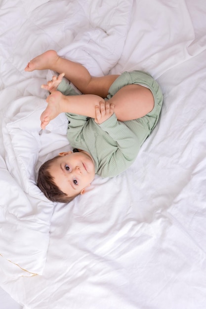 Joli bébé souriant dans un body en coton vert est allongé sur un lit blanc sur son dos vue de dessus