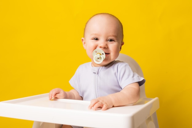 Joli bébé avec une tétine dans sa bouche dans une chaise haute sur fond jaune Espace de nourriture pour bébé pour le texte