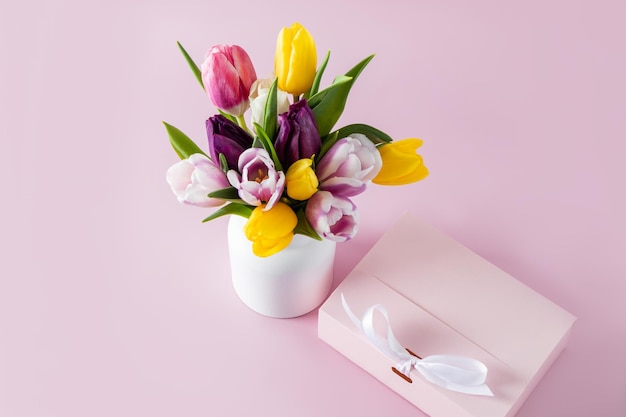 Un joli bouquet de tulipes colorées dans un vase en céramique blanche et une boîte cadeau rose sur une composition festive de fond pastel