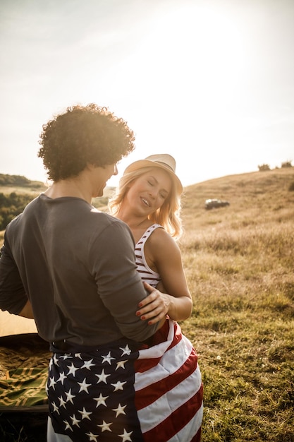 Un joli couple amoureux enveloppé dans un drapeau américain profitant d'une tente au camping.