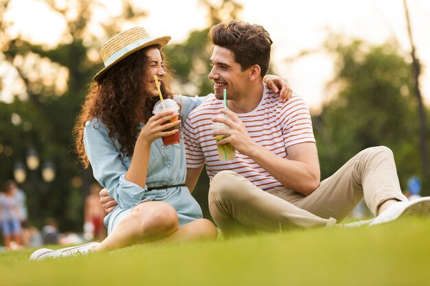 joli couple assis sur l'herbe verte dans le parc et boire des boissons dans des gobelets en plastique