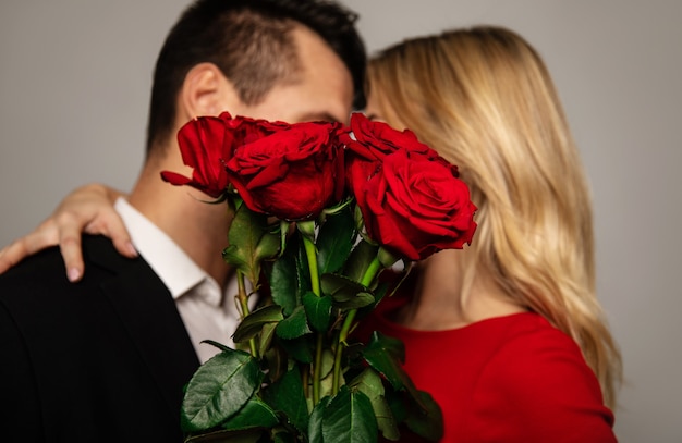 Un joli couple dans des tenues élégantes se cache derrière un beau bouquet de roses rouges tout en s'embrassant et en s'embrassant.