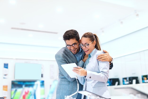 Joli couple multiculturel en tenue de soirée souriant et à la recherche d'une nouvelle tablette à acheter. Intérieur du magasin Tech.