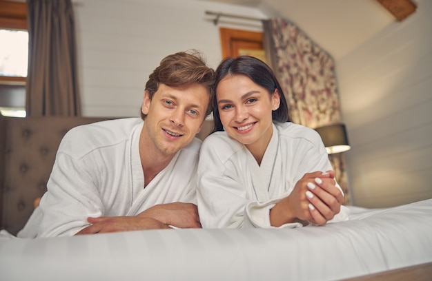 joli couple en peignoirs blancs allongé dans la chambre tout en posant devant l'appareil photo