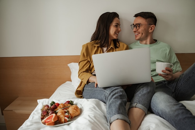 Joli couple regardant un film avec des sentiments romantiques, assis sur le lit avec du café et une assiette de croissants et de fruits frais. Famille heureuse déménageant dans un nouvel appartement.