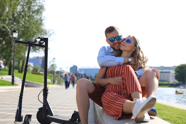 Joli couple en vacances s'amusant à conduire un scooter électrique, faisant une pause dans la conduite, assis au bord de la rivière.