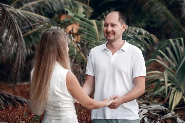 Joli couple vêtu de blanc, main dans la main sur une plage exotique souriante, palmiers ; notion de sentiments.