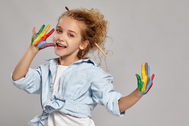 Joli enfant ayant une brosse dans ses cheveux blonds bouclés chics, vêtu d'une chemise bleue et d'un t-shirt blanc. Elle pose avec un bras peint et souriant, sur un fond gris.