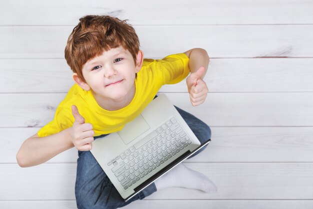 Joli enfant montrant les pouces vers le haut et jouant sur un ordinateur portable sur un sol en stratifié ou en parquet chaud.