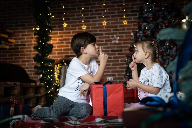 Un joli frère et sœur en pyjama racontant des secrets à côté d'un arbre de Noël dans une pièce décorée