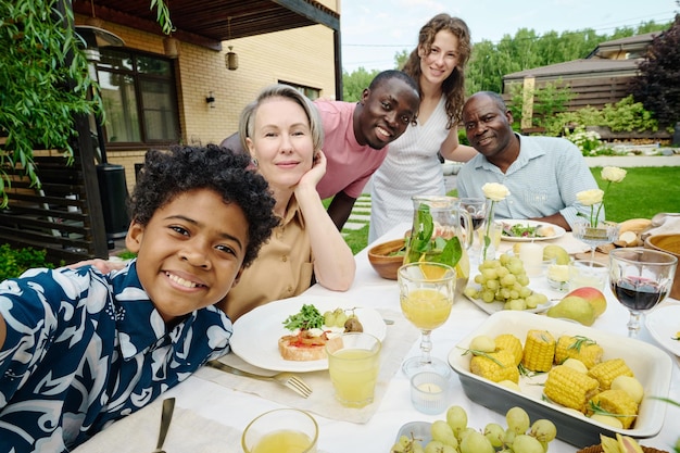 Joli garçon afro-américain en chemise hawaïenne prenant un selfie avec ses parents et ses grands-parents alors qu'il était assis à table pendant le dîner de famille