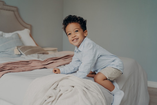 Joli garçon afro-américain heureux jouant et s'amusant sur le lit après son retour de la maternelle, jouant et souriant tout en passant du temps dans la chambre. Concept d'enfance insouciante