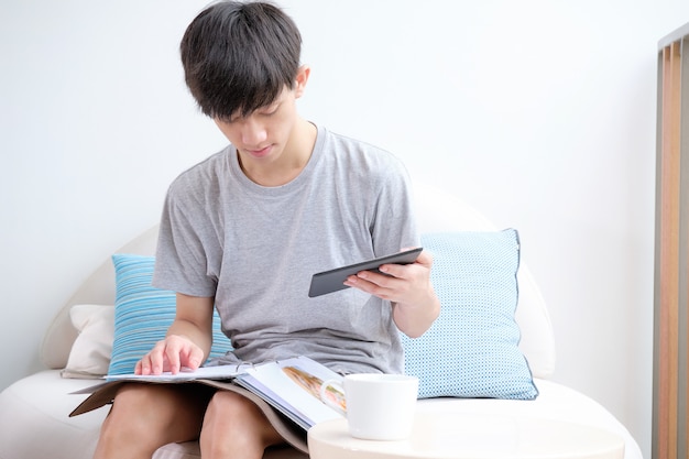 Joli garçon assis sur un canapé-lit tandis que sa main tenant un livre et une tablette pour faire du shopping en ligne.