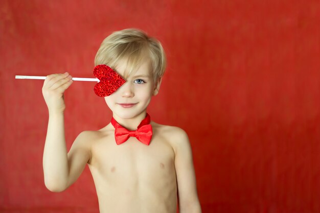 Joli garçon Cupidon avec un triple sourire, noeud papillon et flèche coeur isolé sur mur rouge. Concept de la Saint-Valentin.