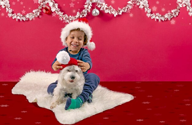 Photo joli garçon jouant et riant avec son petit chien mettant un bonnet de noel dans une décoration de noël