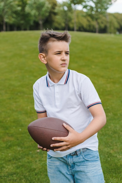 Joli garçon en polo tenant un ballon de rugby et regardant loin dans le parc